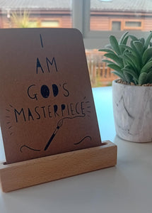 Brown sign with 'I AM GOD'S MASTERPIECE' text on a wooden stand, next to a potted plant.