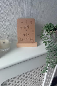 Small wooden sign with 'I am a new creation' text on a white surface with a plant and candle in the background.