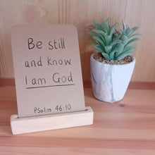 Load image into Gallery viewer, Note with &#39;Be still and know I am God&#39; on a wooden stand next to a small plant on a wooden surface.
